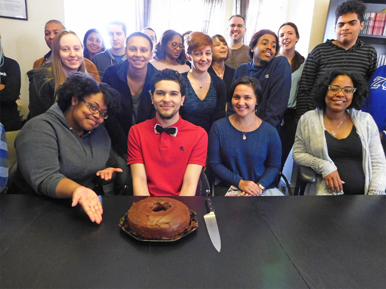 Brandon (wearing a red shirt) sits at a long black table with a chocolate cake while surrounded by his friends, and fellow Hopeworks interns in the huddle room at the former at the former Hopeworks nonprofit in Camden, New Jersey.